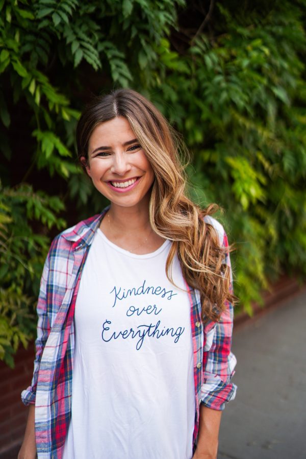 Picture of Becky Epstein wearing "kindness over everything" shirt with a flannel shirt and a green leaf background.